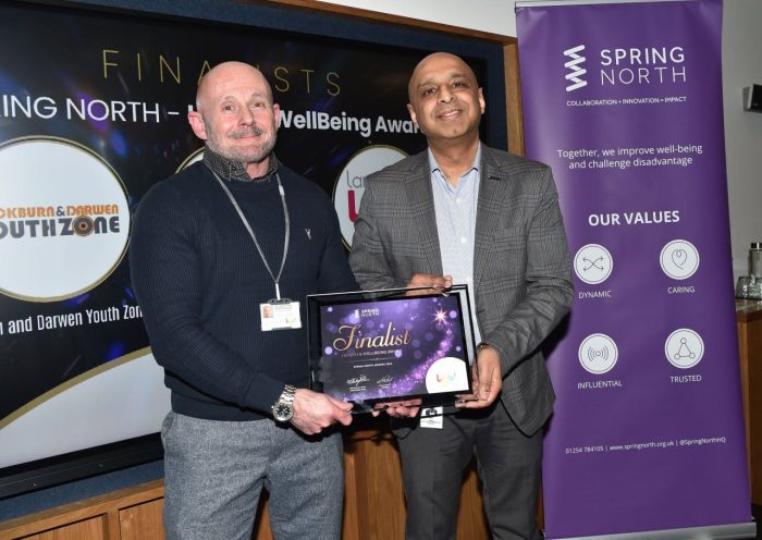 Two men stood in front of a purple Spring North pop-up banner, and a sign saying Finalist - Health and Wellbeing Award. Lewis, Lancashire LGBT'S Chief Executive Officer, is stood to the left of a man in a grey blazer handing Lewis a framed certificate that says Finalist. Lewis is wearing a navy blue jumper and grey pants.