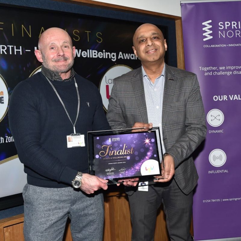 Two men stood in front of a purple Spring North pop-up banner, and a sign saying Finalist - Health and Wellbeing Award. Lewis, Lancashire LGBT'S Chief Executive Officer, is stood to the left of a man in a grey blazer handing Lewis a framed certificate that says Finalist. Lewis is wearing a navy blue jumper and grey pants.