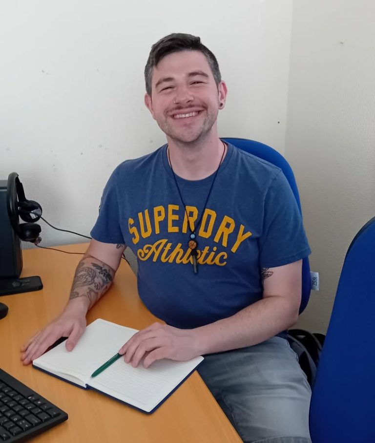 Jake, a young man in his early 30s with brown eyes and brown hair with hints of grey on both sides of his head, is sat on a blue desk chair, and his arms are resting on a brown computer desk. In front of Jake is an open blank notebook and a green pen resting on the notebook's pages. Jake is wearing a blue shirt, grey shorts, and a crystal necklace. Jake has several tattoos on his right arm, has a silver circular piercing in his right nostril, and is smiling gleefuly.