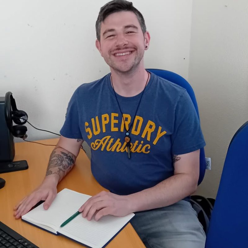Jake, a young man in his early 30s with brown eyes and brown hair with hints of grey on both sides of his head, is sat on a blue desk chair, and his arms are resting on a brown computer desk. In front of Jake is an open blank notebook and a green pen resting on the notebook's pages. Jake is wearing a blue shirt, grey shorts, and a crystal necklace. Jake has several tattoos on his right arm, has a silver circular piercing in his right nostril, and is smiling gleefuly.