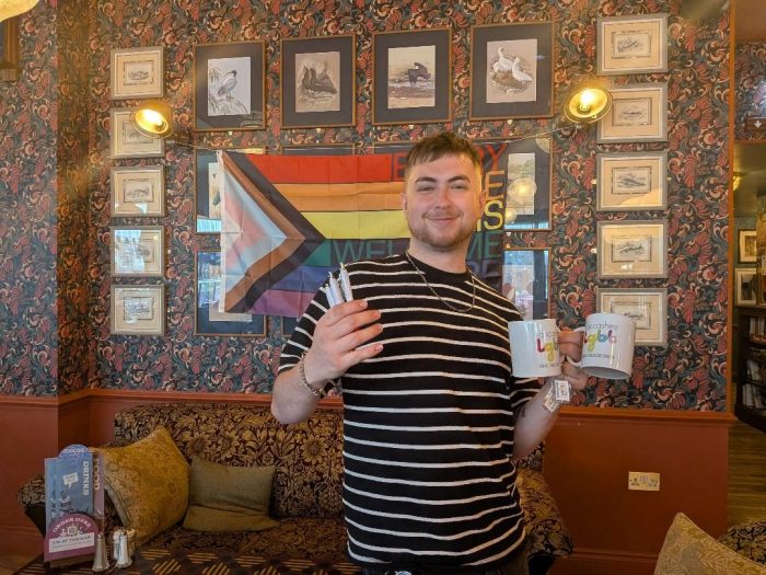 Connor, who works at Argento Lounge, is stood smiling in front of a Pride progress flag pinned up against a wall with framed pictures around the flag. Connor is wearing a black and white stripped t-shirt. In his left hand, he is holding two white mugs with the Lancashire LGBT logo on it, and in his right hand he is holding three Lancashire LGBT pens.