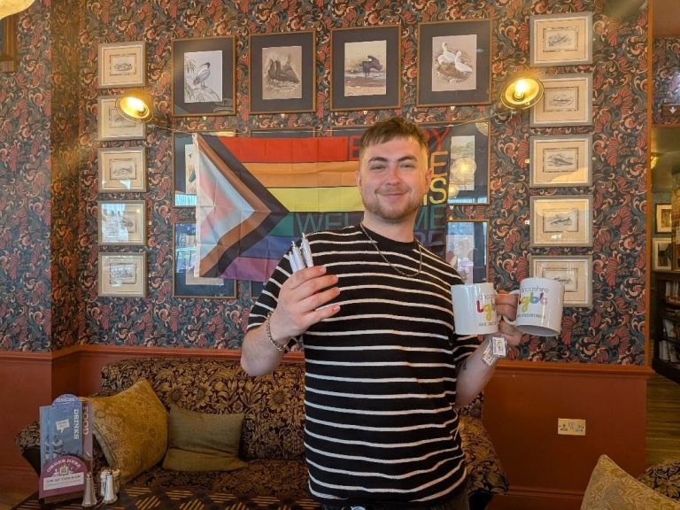 Connor, who works at Argento Lounge, is stood smiling in front of a Pride progress flag pinned up against a wall with framed pictures around the flag. Connor is wearing a black and white stripped t-shirt. In his left hand, he is holding two white mugs with the Lancashire LGBT logo on it, and in his right hand he is holding three Lancashire LGBT pens.