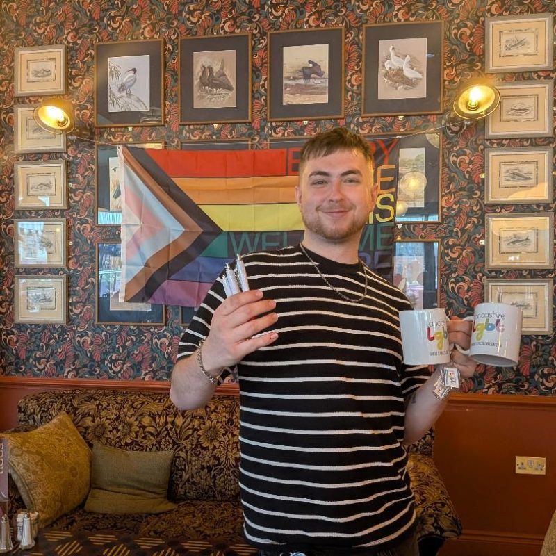 Connor, who works at Argento Lounge, is stood smiling in front of a Pride progress flag pinned up against a wall with framed pictures around the flag. Connor is wearing a black and white stripped t-shirt. In his left hand, he is holding two white mugs with the Lancashire LGBT logo on it, and in his right hand he is holding three Lancashire LGBT pens.