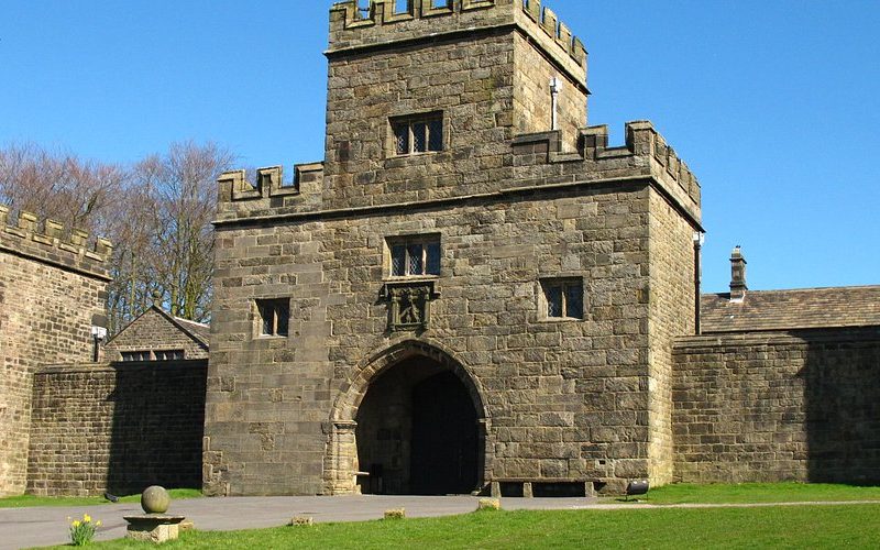A picture of Hoghton Tower gatehouse and walls, set against a bright blue sky. There is a grassy lawn in front of the wall with a stone pathway next to it.